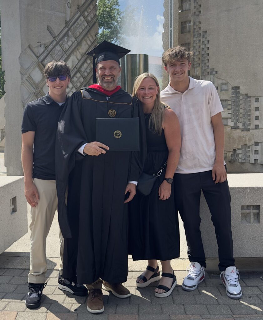 Jason Bristow poses with his family on his graduation day. He is wearing Purdue graduation regalia and standing in front of the Purdue arch.