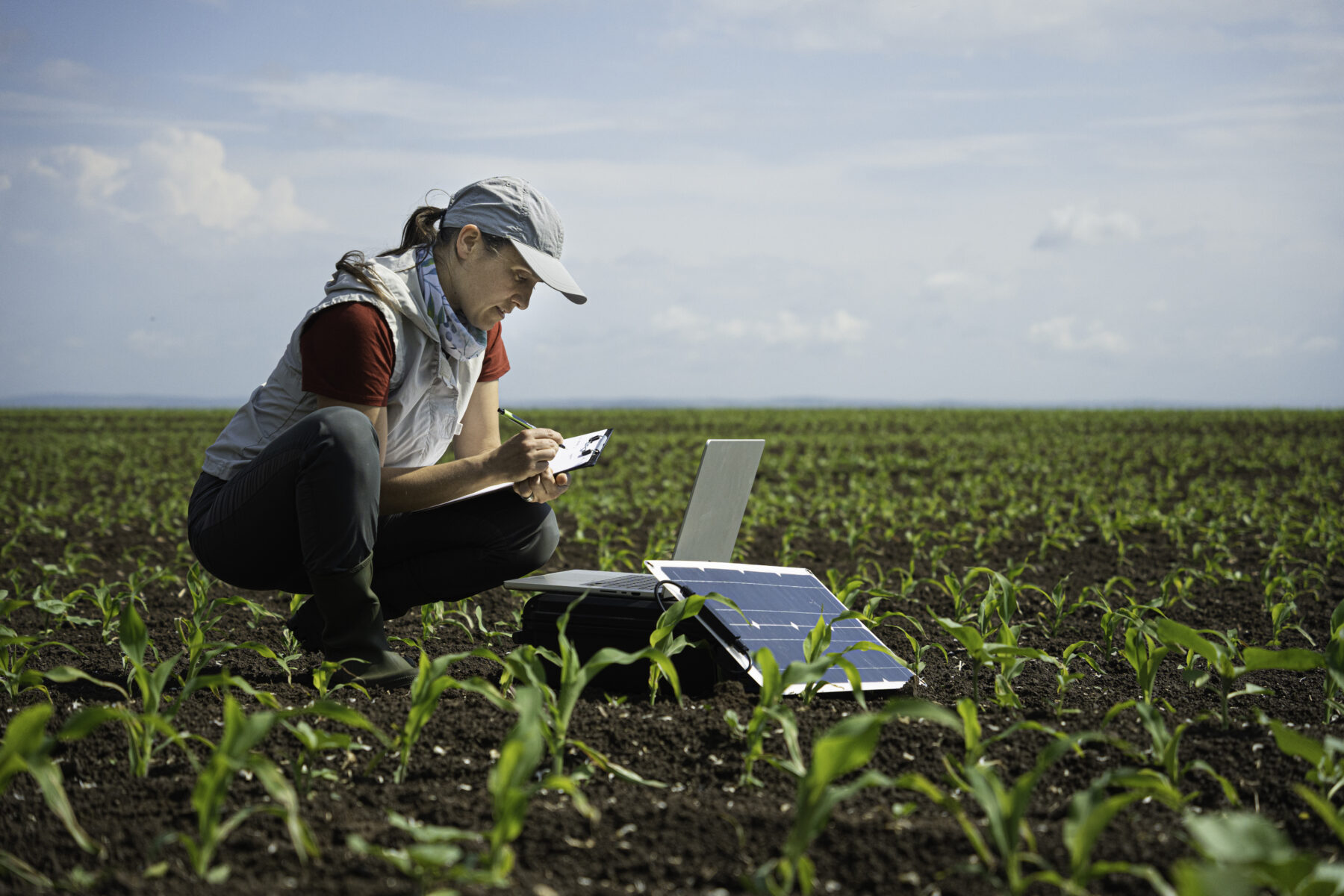 A woman sits in a field taking notes on a notepad. A laptop and solar panel sit in front of her.