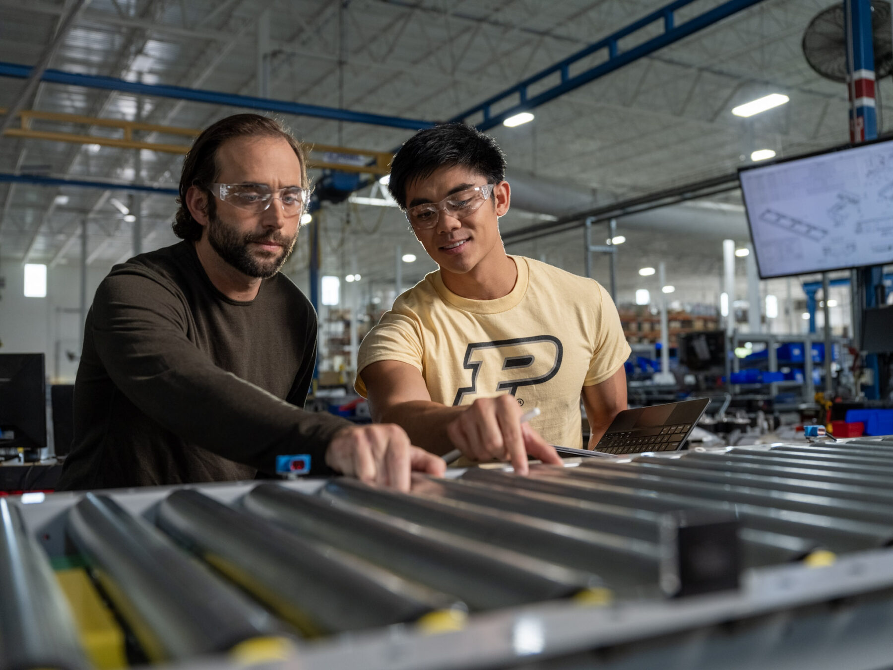 Two men work together on a product line. The man on the right is wearing a gold shirt with the Purdue 