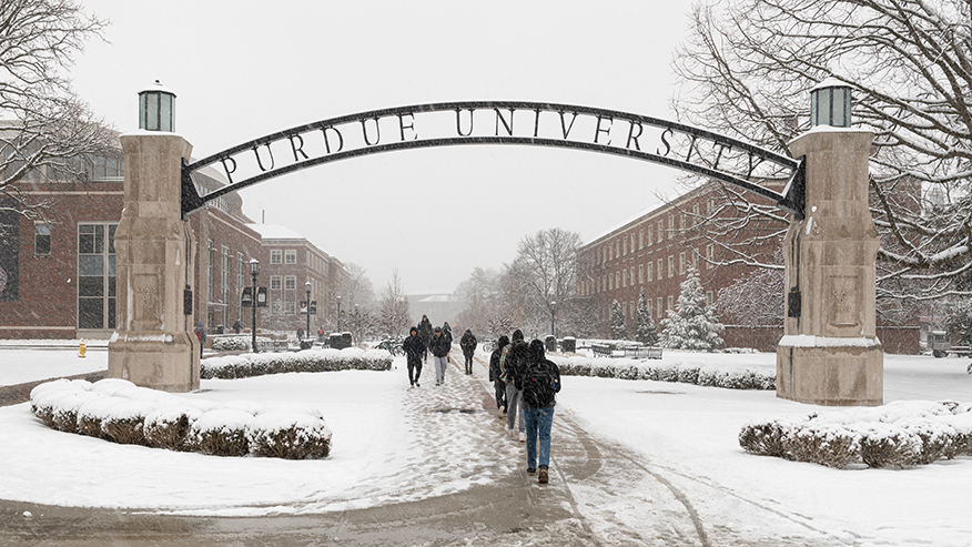 Purdue students walk under a campus archway on a snowy day. The archway says 