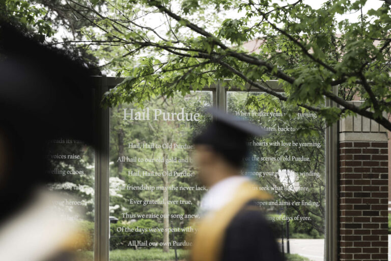 A Purdue student in graduation attire walks past a "Hail Purdue" placard on campus.