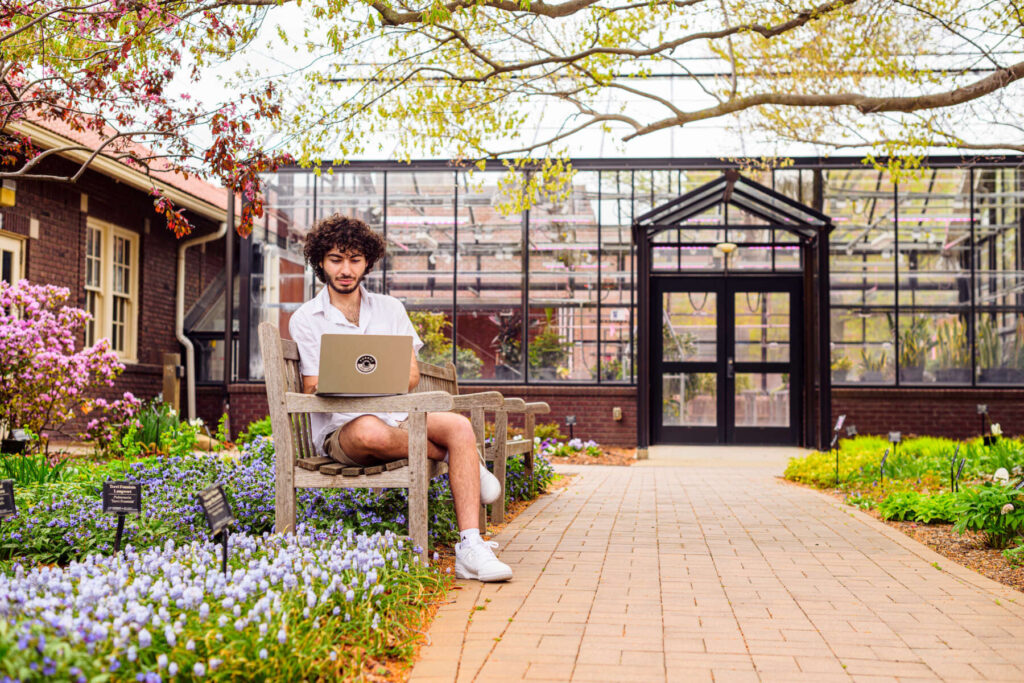 student on a laptop sitting near a greenhouse