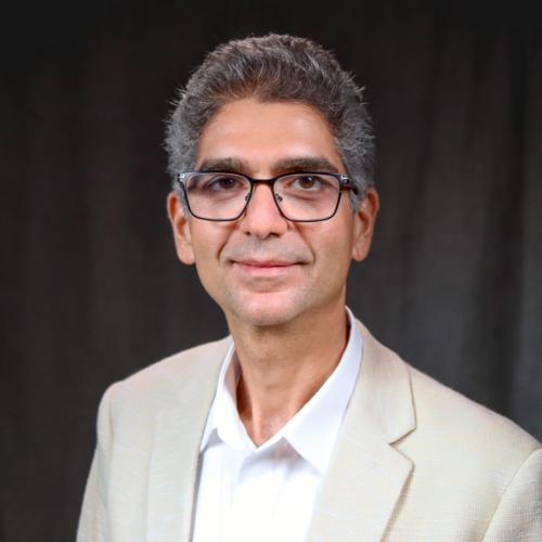 Rami Ghannam smiles while looking at the camera. He is wearing a tan and white suit. 