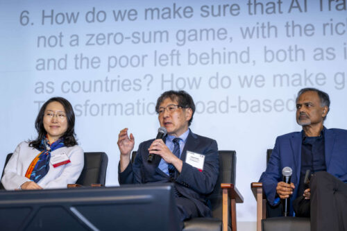 (From left to right): Yorie Nakahira, associate professor of electrical and computer engineering at Carnegie Mellon University; Hideaki Ozawa, chief technology officer at NTT Research, Inc.; and Kaushik Roy, Edward G. Tiedemann Jr. Distinguished Professor of electrical and computer engineering at Purdue University. A group of three panelists address a crowd at Purdue University. A question about AI is projected behind them.
