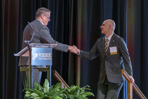 Purdue Senior Vice President for Partnerships and Online Dimitrios Peroulis with keynote speaker Mike Dolan. Mike Dolan and Dimitrios Peroulis shake hands behind a podium with the Purdue University logo on it.
