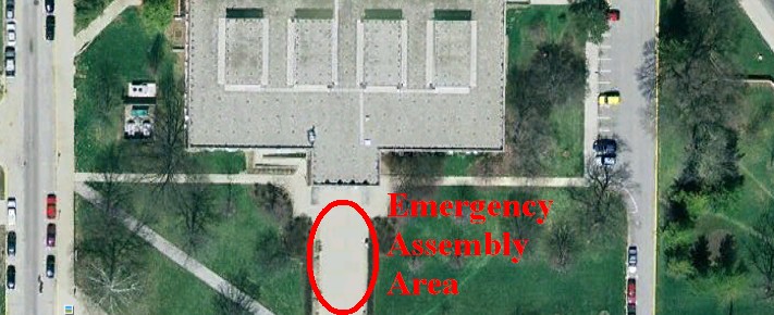 “Aerial photograph of Ward L. Lambert Gymnasium surrounded by parking lots, driveways, and clusters of trees. Cars are parked in several rows around the property. In front of the building, a concrete sidewalk area is outlined with a red oval and labeled ‘Emergency Assembly Area’ in red text, indicating the designated Emergency Assembly Area (EAA) gathering point during an evacuation.”