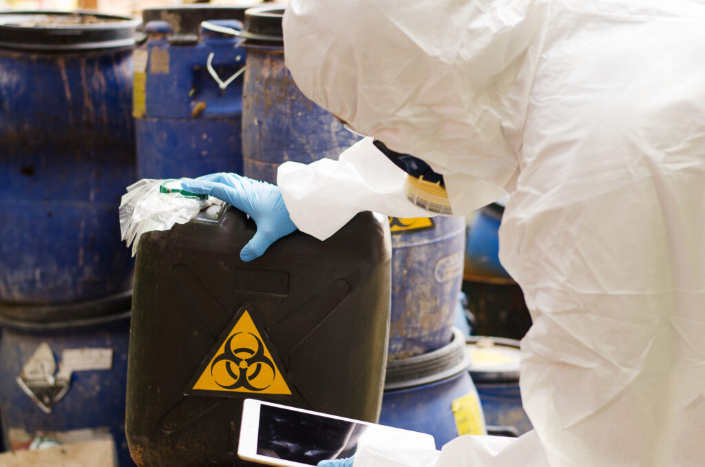 A person in a hazmat suit surrounded by barrels with a hazardous waste symbol on them.
