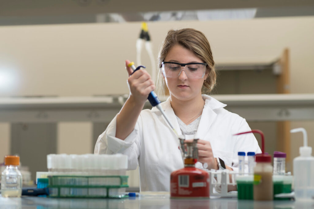 A scientist in a lab coat with safety glasses on injecting something into a vial.