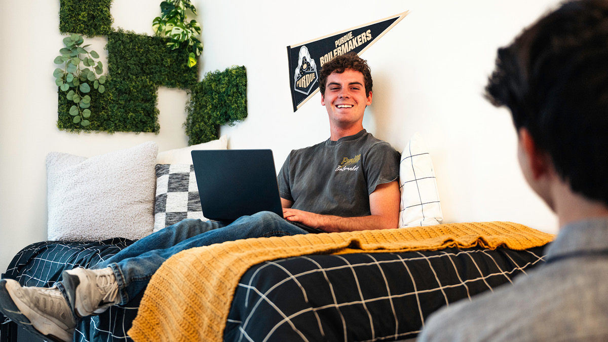 A young male student sitting on a bed with a laptop speaking to a friend.
