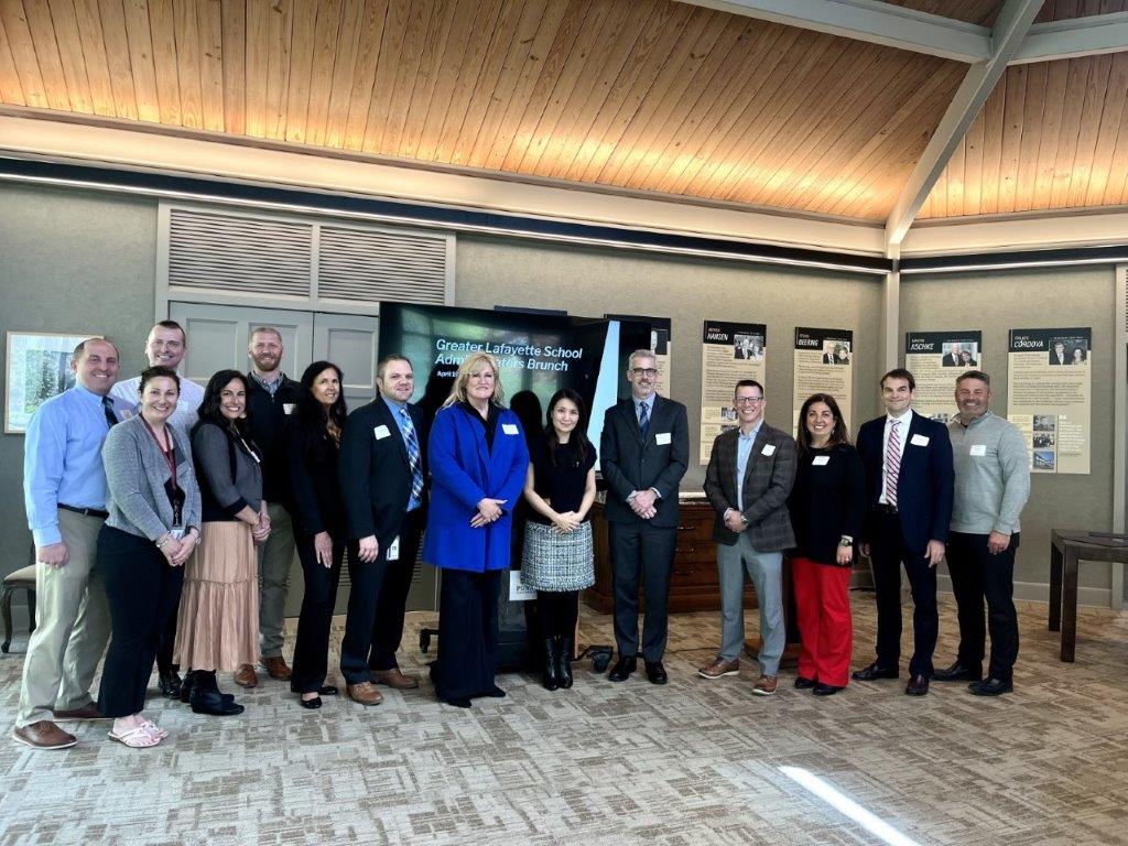 A group of professionals posing for a photo in front of a screen at the Greater Lafayette School Administrators Brunch.