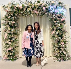 Two women smiling in front of a large floral arch with an "Oh Baby" neon sign.