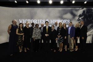 A group of people posing for a photo on a stage in front of a "Victories & Heroes" backdrop.