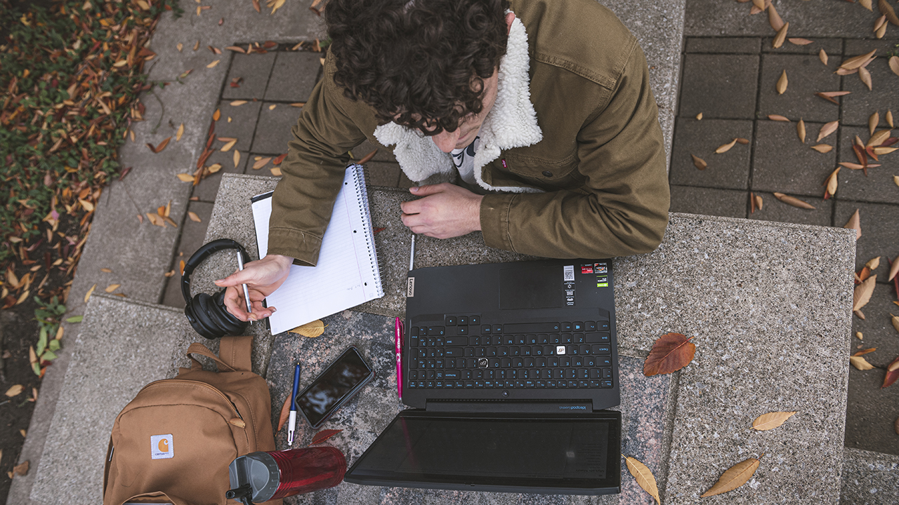 A student looks at his laptop as he writes notes working at a table outside on a fall day