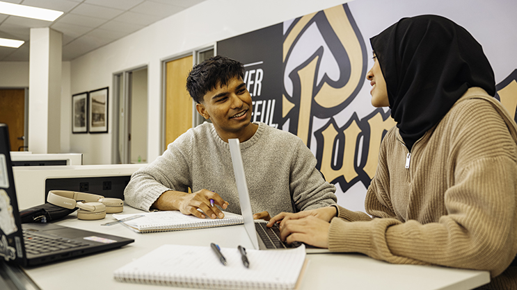 Two students talk as they work side-by-side at a workstation