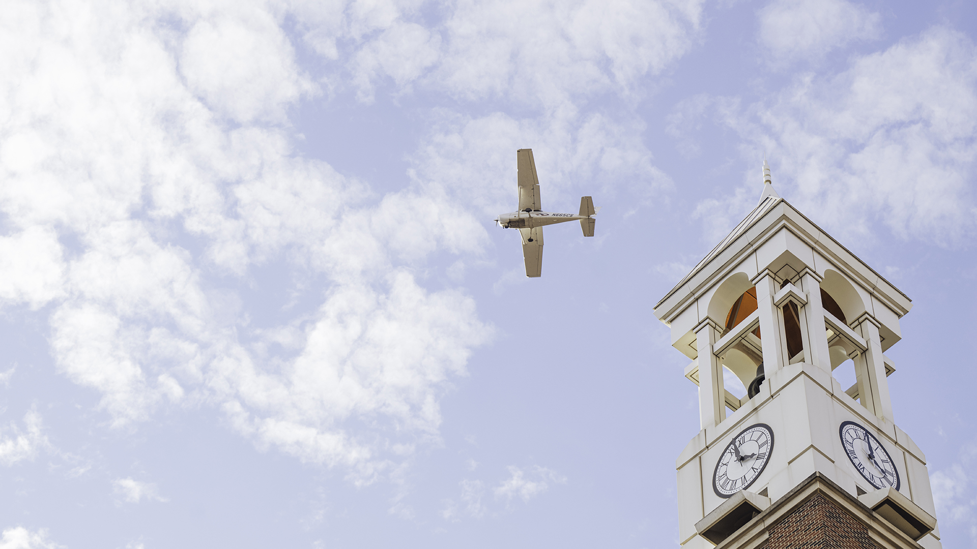 A Purdue airplane flies above the Bell Tower against a blue sky