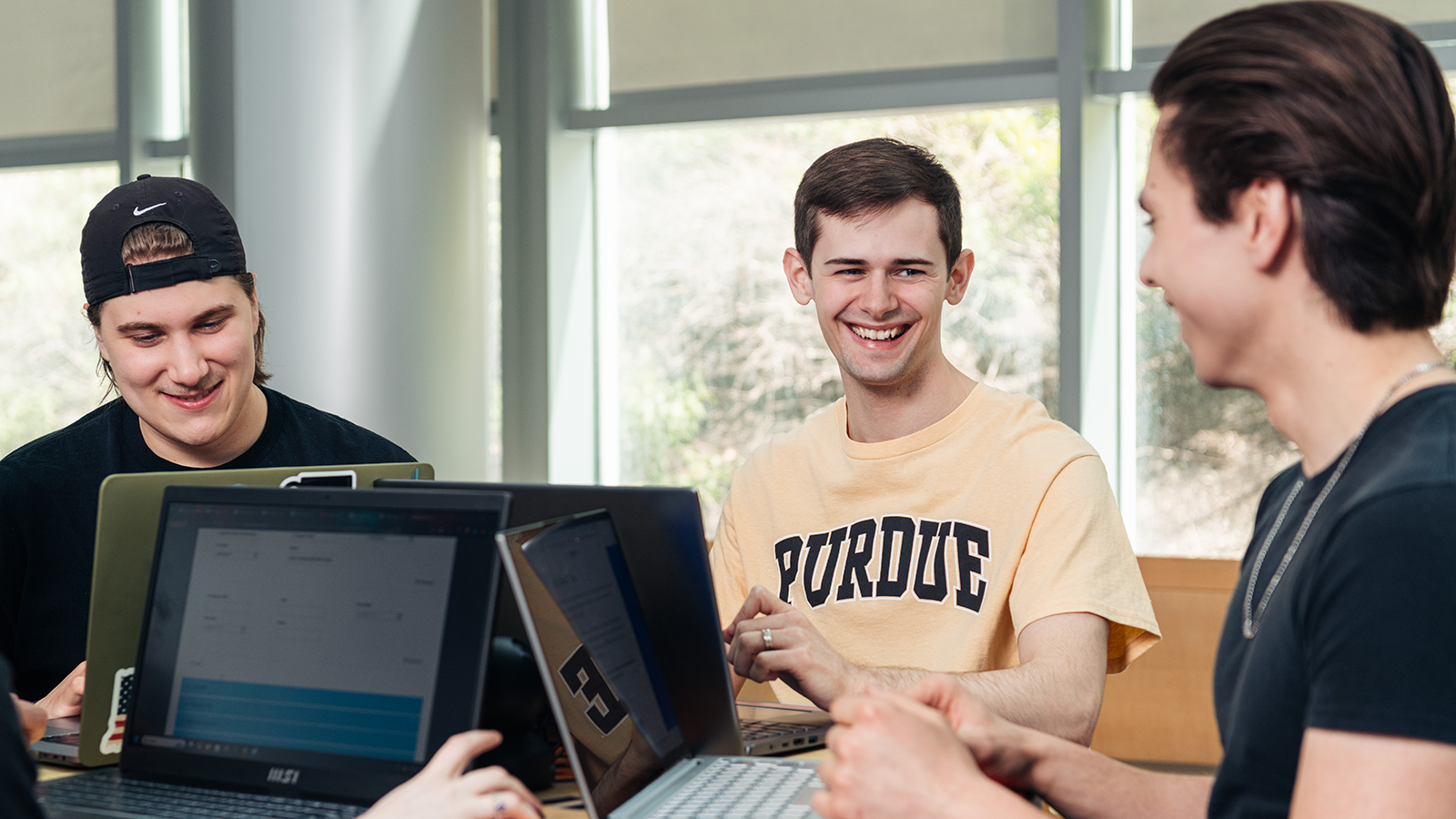 Three students sitting at a table facing each other with their laptops