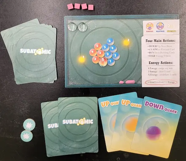 Photo 3. The Player Mat includes a key at the top right to help players remember which group of cards can purchase protons, neutrons, or electrons. Pictured here, a player used 2 Up Quark cards and 1 Down Quark card to purchase 1 proton (red) on the Player Mat. A Particle Marker was placed on a Proton to help the player keep track of the Proton they purchased with their cards.