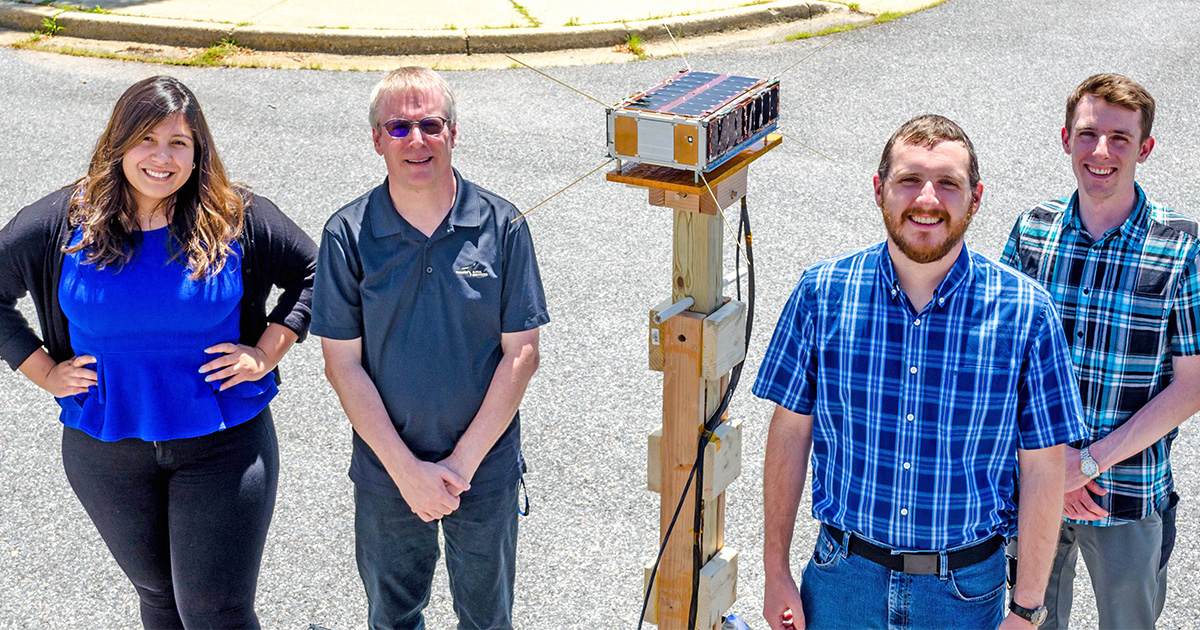 Purdue students and faculty member Jim Garrison stand with the SNOOPI cubesat mounted to a wooden stand.