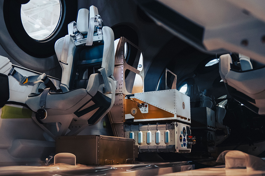 Two metal boxes holding automated experiments mounted to the floor behind a passenger seat inside the cabin of Virgin Galactic's Unity spacecraft