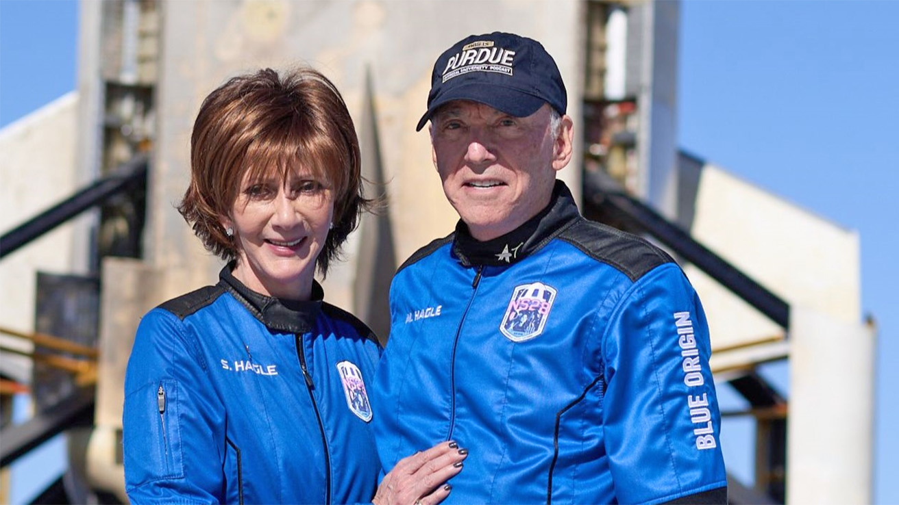 Sharon and Marc Hagle standing in blue astronaut jump suits near a rocket on a launchpad.