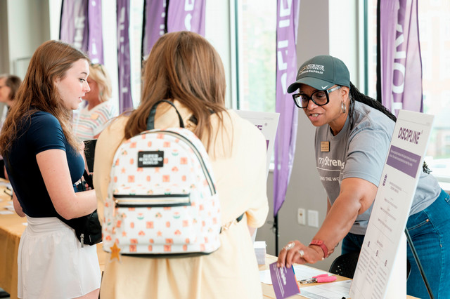 A staff member interacts with students over a table at an orientation event. 
