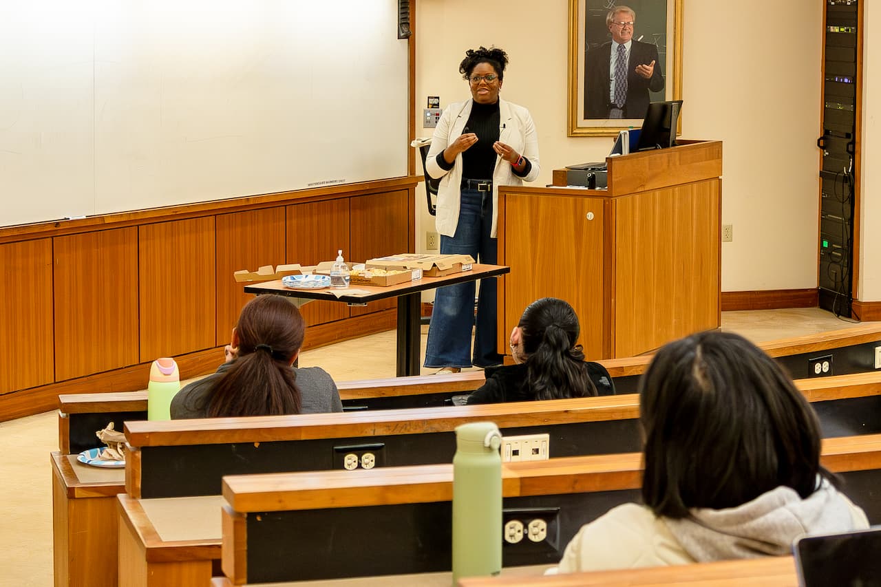 A woman stands in front of a classroom lecturing with rows of students facing her.