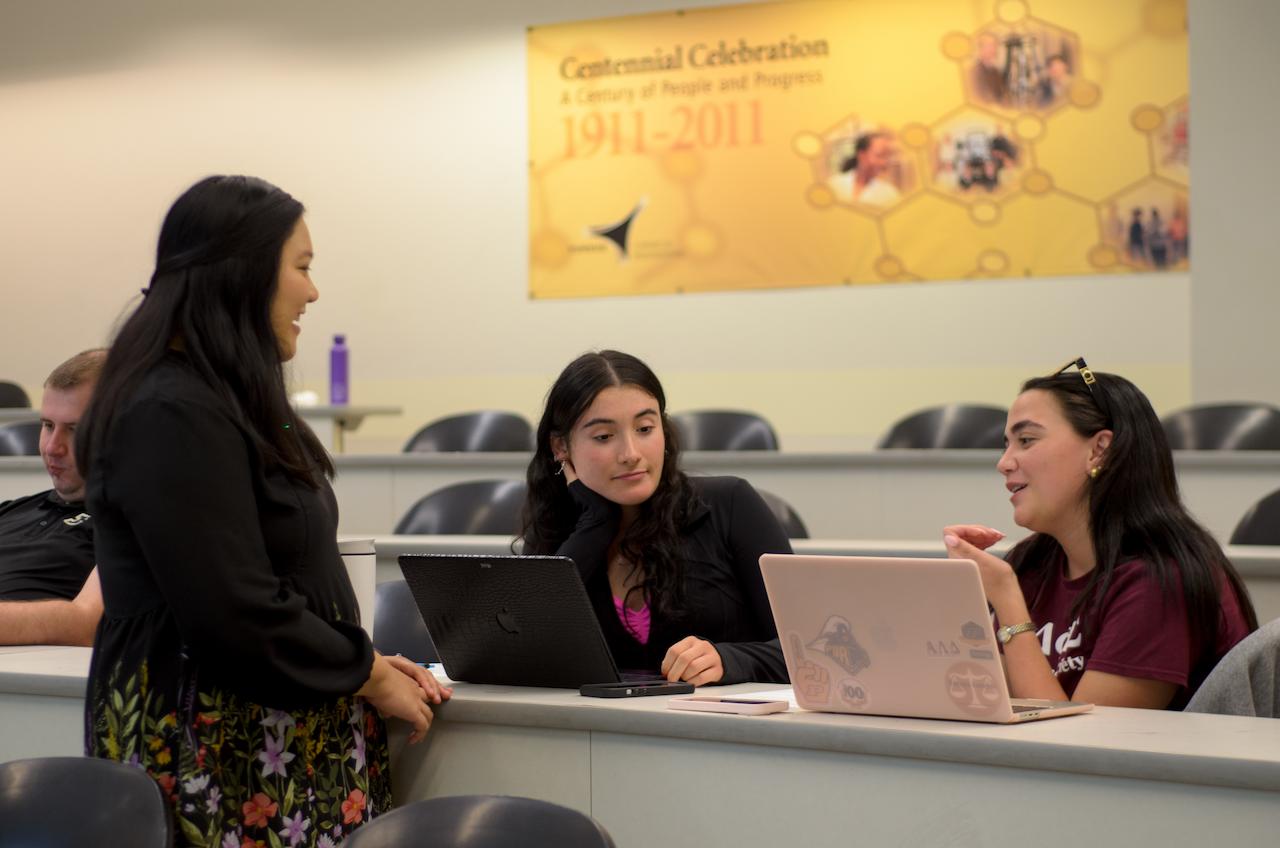 Three female students gather around laptops in a classroom discussing content on the screens.