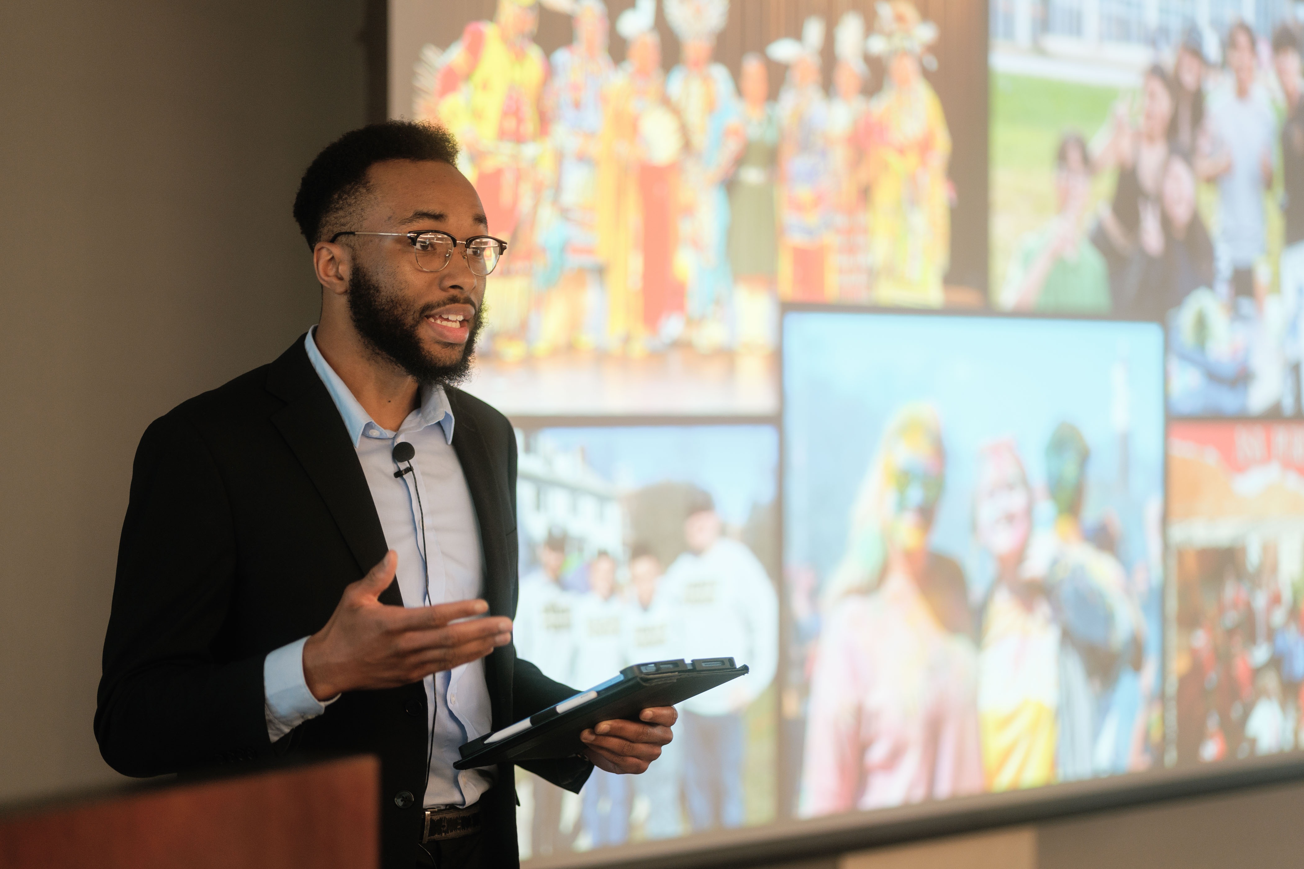 A man makes a presentation with a large screen full of colorful photos behind him.